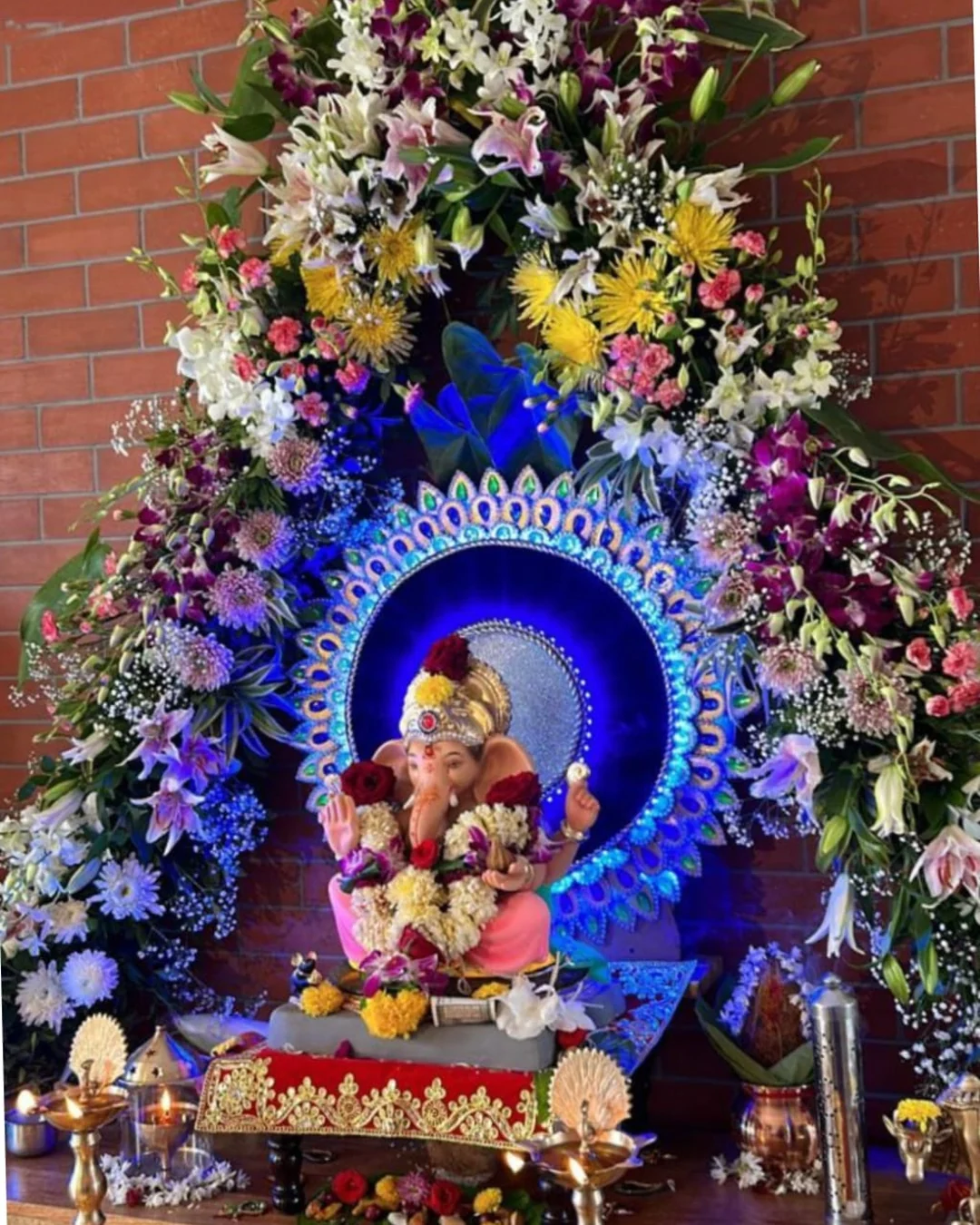 Ganpati idol framed by a blue peacock halo and lily-orchid arch, brass diyas alight